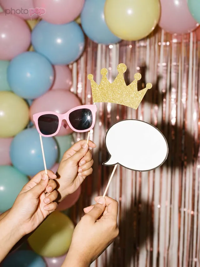 Hands holding pink sunglasses, a gold glitter crown and a blank speech-bubble prop in front of pastel balloons and a rose-gold tinsel curtain