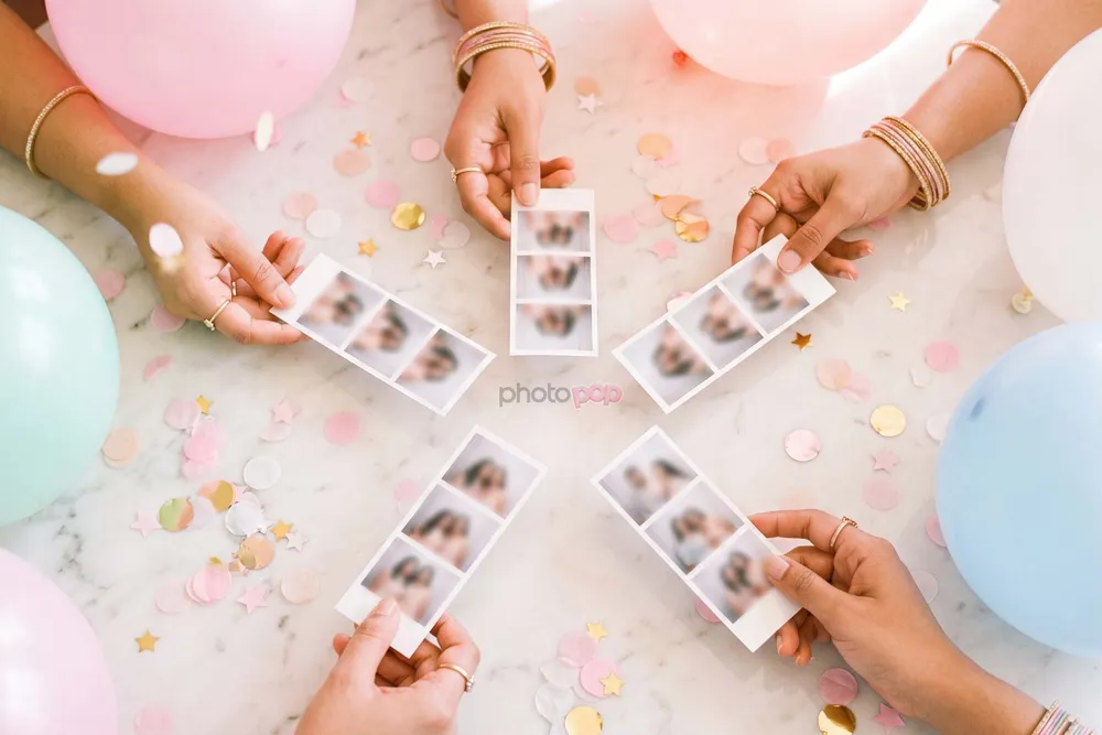 Five hands holding photo strips in a circle on a marble table scattered with pastel confetti and balloons