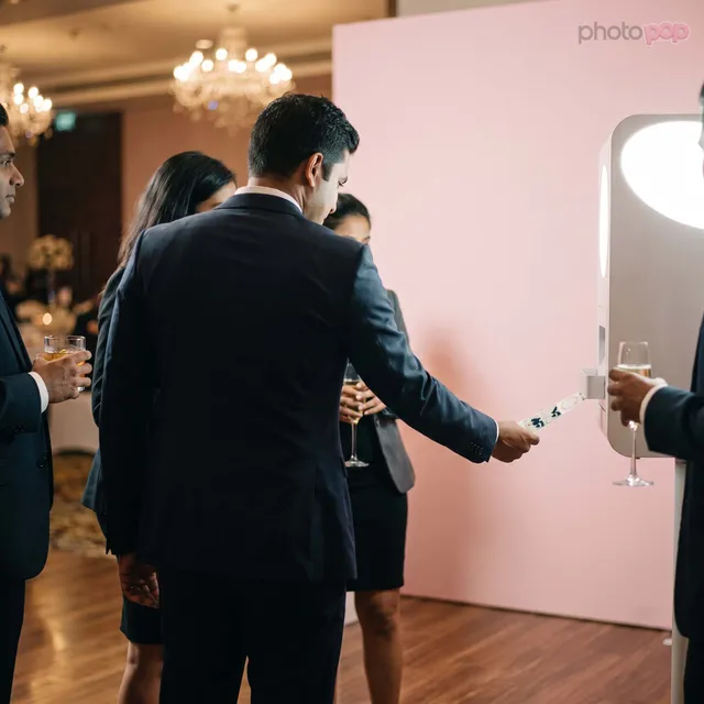 Guests in suits with drinks gathered at a photo booth against a pink backdrop in a chandeliered ballroom, one collecting a print