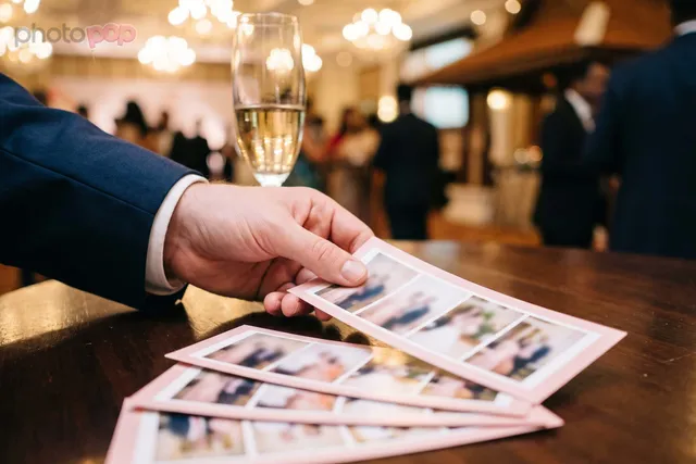 A hand picking up photo strips with blush-pink borders from a wooden table, a champagne glass and ballroom lights behind