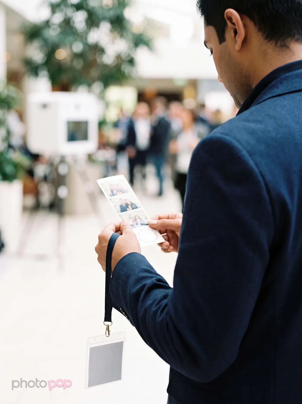 A conference guest with a lanyard studying a photo strip, a photo booth blurred in the bright atrium behind