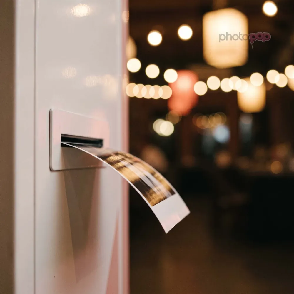 A glossy photo print curling out of a white photo booth's print slot, warm festival lights blurred behind