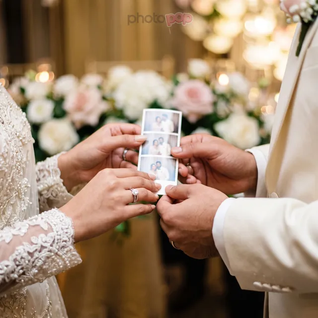 A bride and groom's hands holding a fresh three-frame photo strip, with white and pink roses behind them