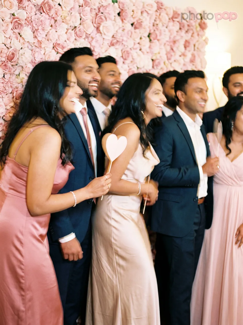 A laughing bridal party in blush dresses and navy suits posing with heart-shaped props in front of a pink rose wall