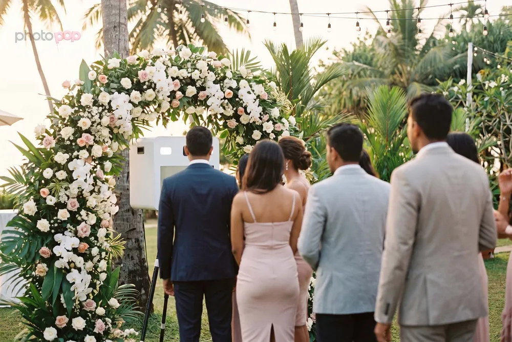 Guests queuing at a photo booth framed by a white and blush floral arch in a tropical garden at golden hour